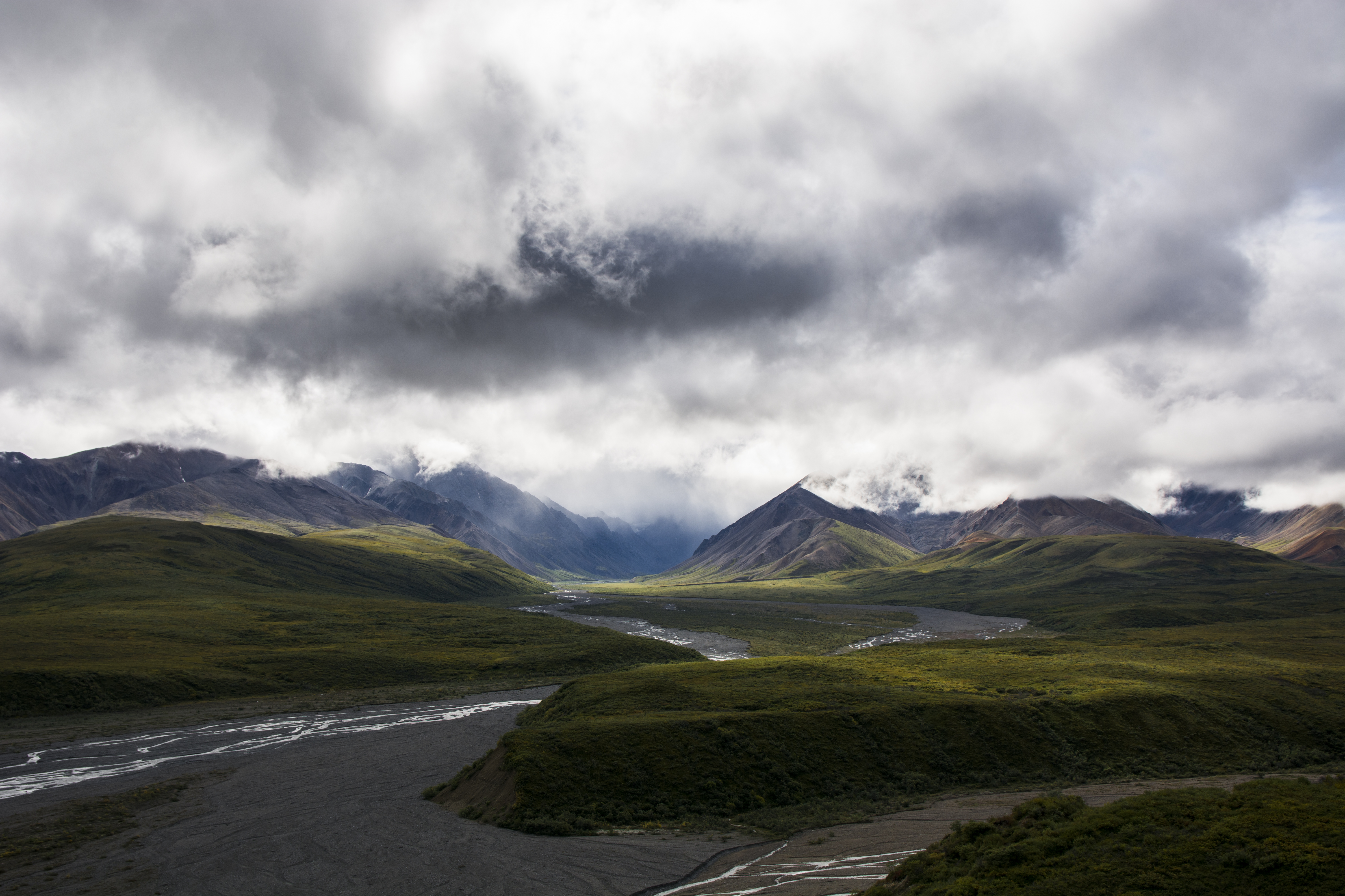 River valleys and green tundra lead up into rocky mountains. The tops of the mountains are shrouded in gray clouds.