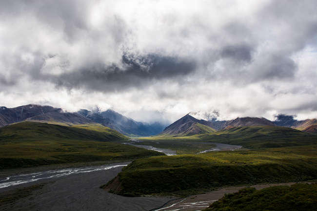 River valleys and green tundra lead up into rocky mountains. The tops of the mountains are shrouded in gray clouds.