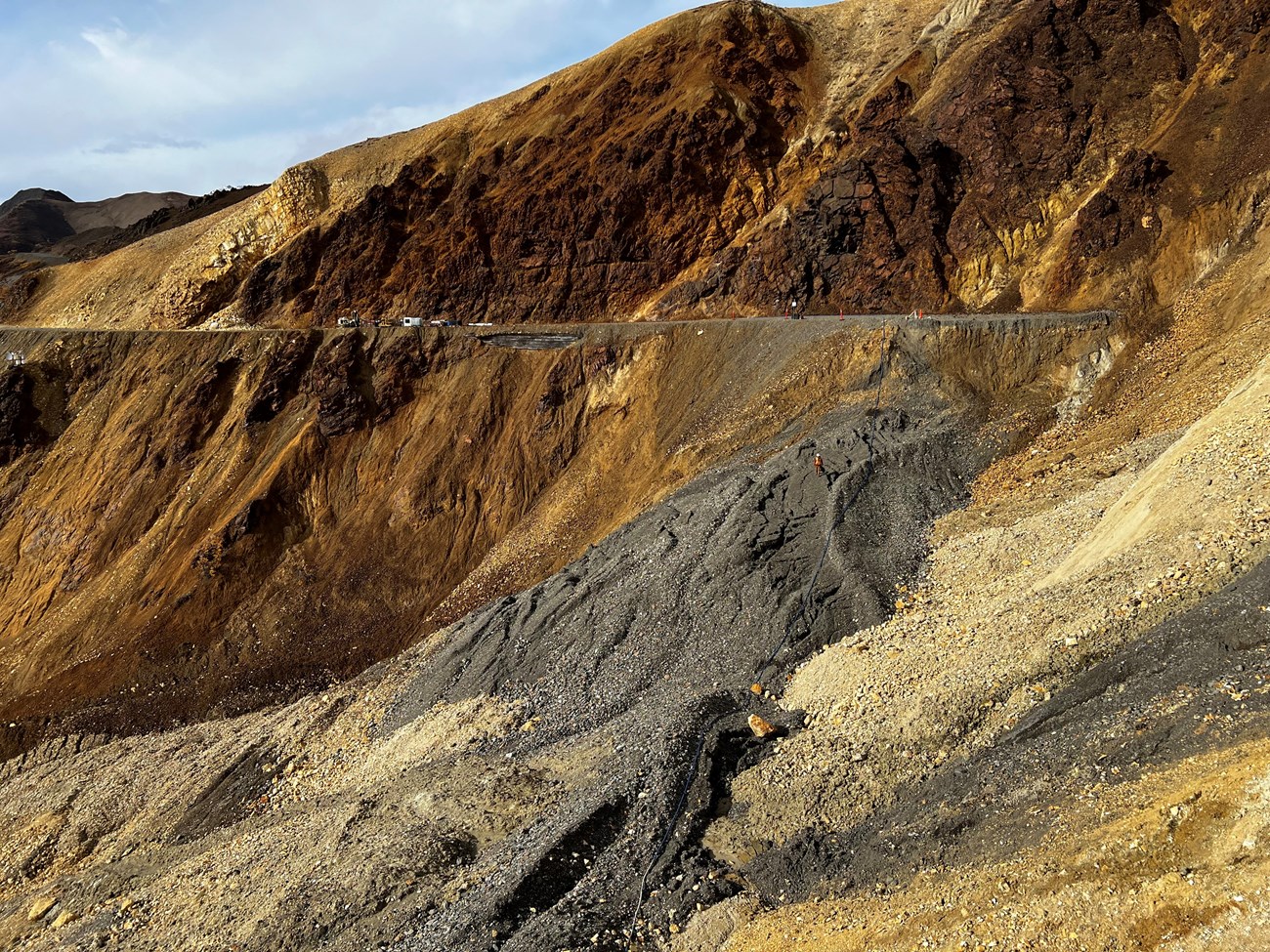A mountainside of bare orange, yellow, brown, and gray rock. On the far side of the slope, a flat road is cut into the mountain, then suddenly drops away and piles of slumping rock can be seen below the road. Two people work with equipment on the rock.