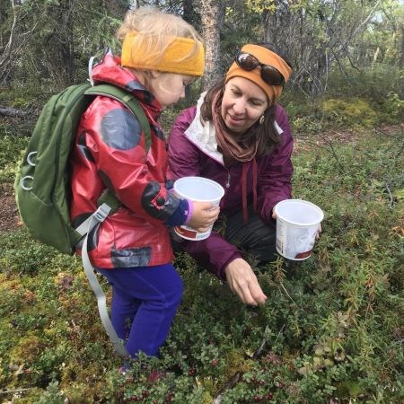 A woman and a toddler in a forest are holding empty containers and picking low-bush cranberries.