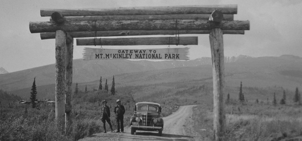 two people stand next to a car on a dirt road under a sign for Mt. McKinley National Park