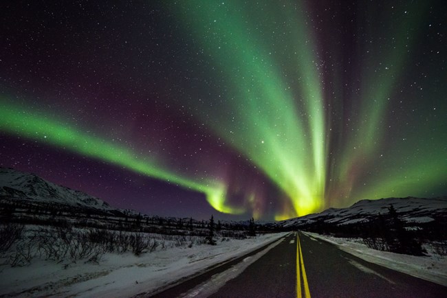 Bright green and purple aurora streaks across the sky, a dark road and snow-covered mountains are in the foreground.