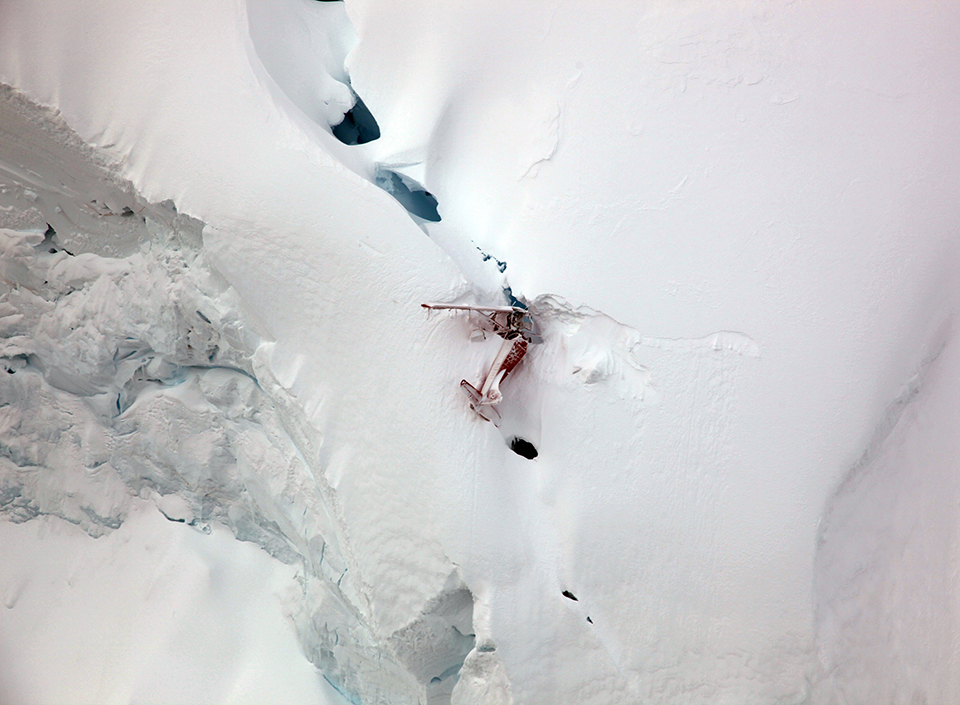 Image of aircraft crash taken from above. The plane appears severely damaged and embedded into the snow. The aircraft is on the side of a steep drop of a glacier wall.