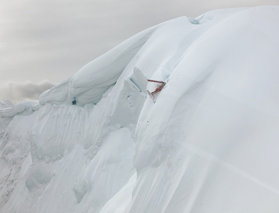 Image of aircraft taken from an oblique angle that shows a steep slope with a plane embedded into the snow. The plane appears to be severely damaged.