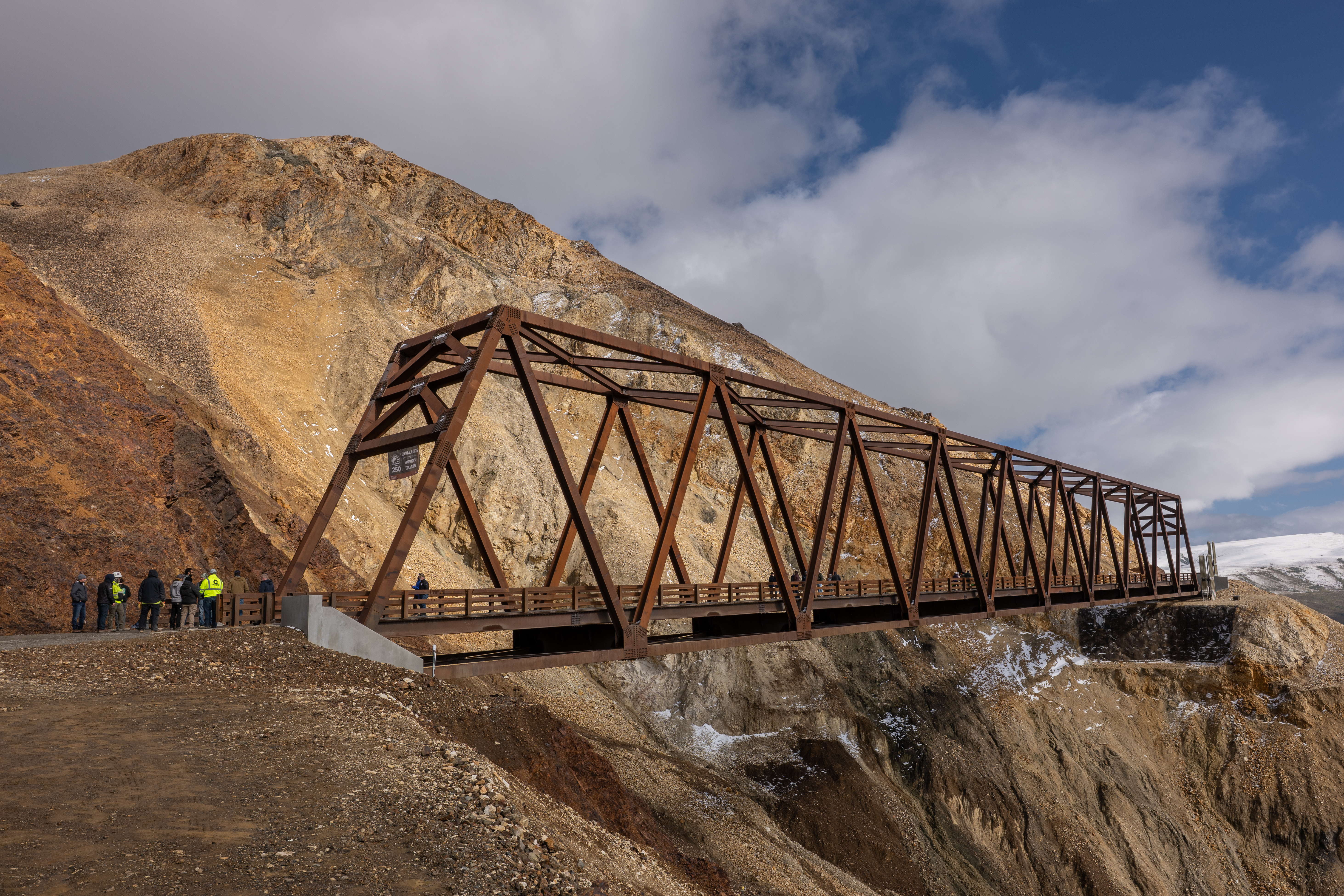 A red trestle bridge expands over a rocky slope of pale rocks with a cloudy blue sky in the background.