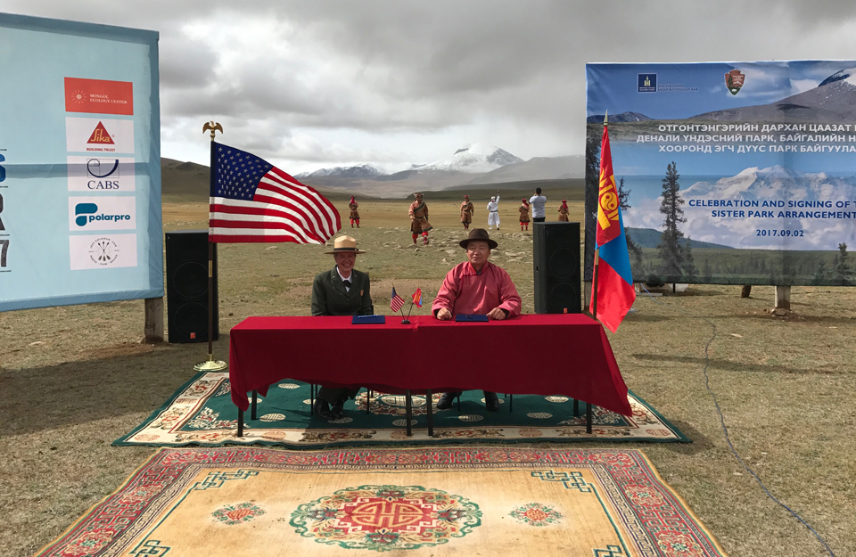 a park ranger and a man in traditional mongolian attire sit at a table outside in front of flags and posters