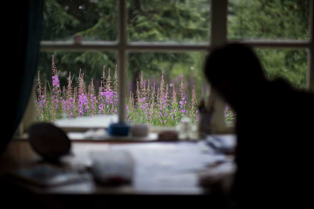 silhouette of a man drawing at a desk in front of a window with a view of pink flowers outside