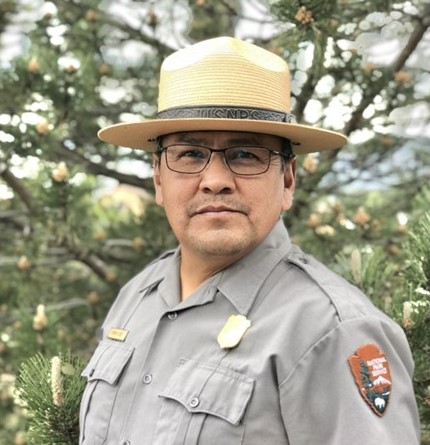 A headshot of a man wearing a ranger uniform, standing outside in front of a tree.