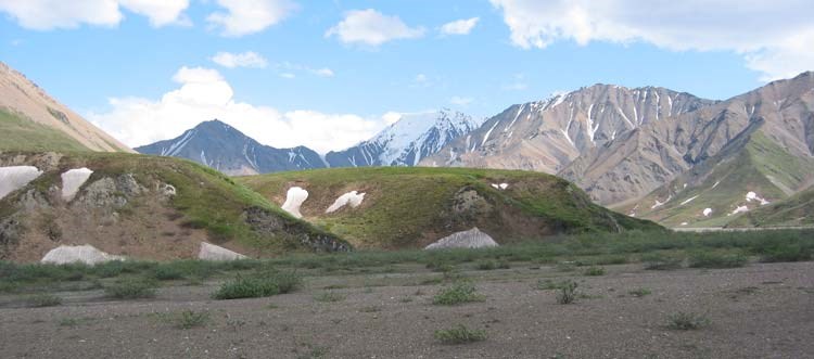 a gravel plain leading up to green hills, in front of taller, snow-dotted mountains