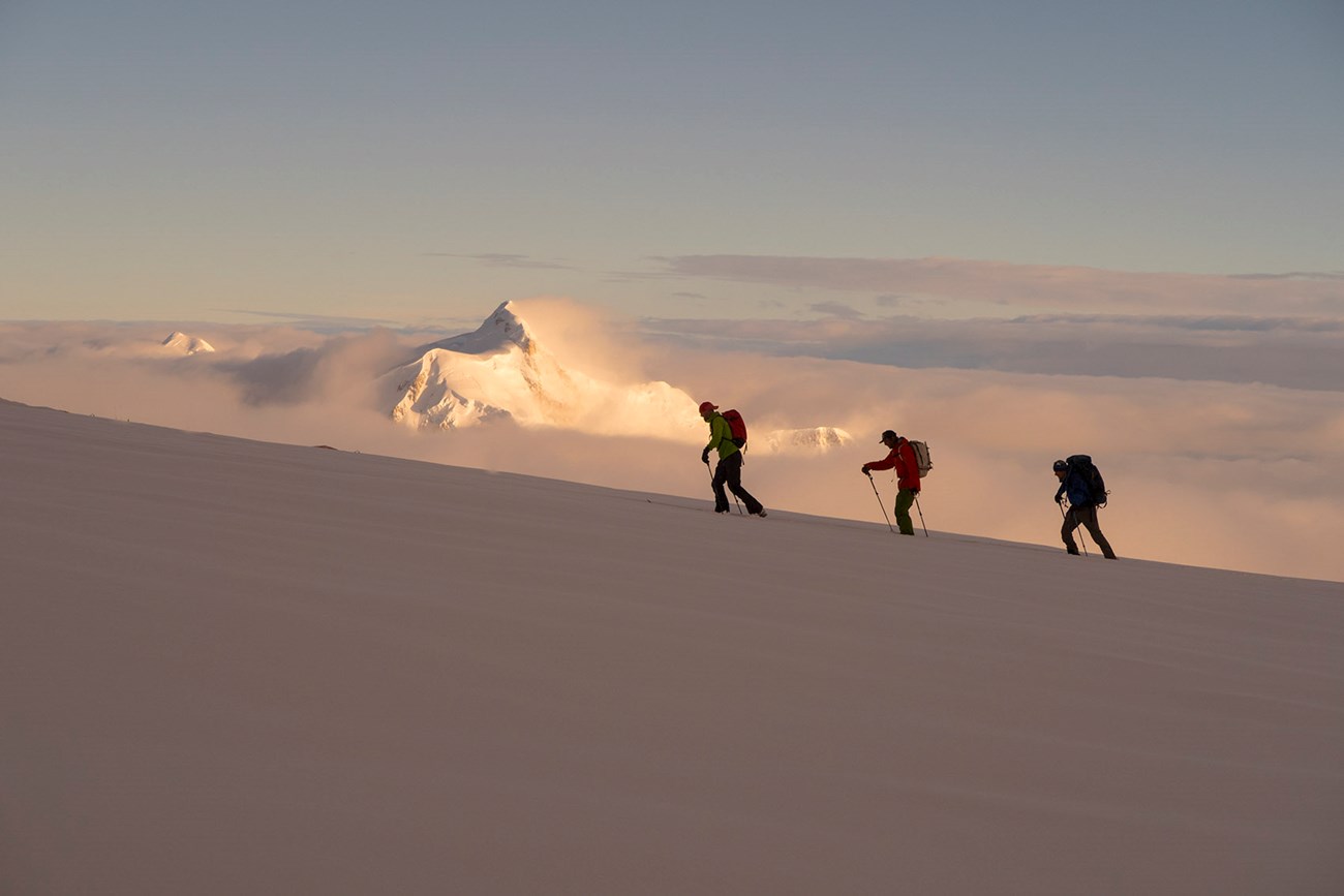 Mountaineers ascend a shadowy slope, with a glowing snowy peak in the distance