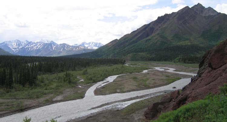 a river running through forest, past a tall mountain, other mountains in the distance