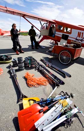 a small plane on a runway with two men and piles of skis and other equipment nearby