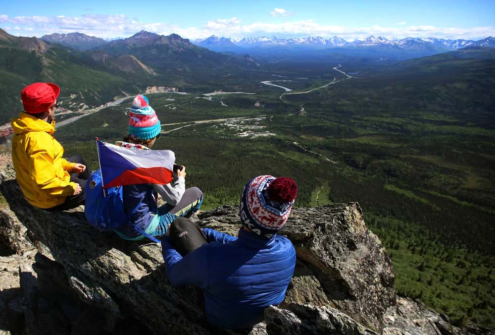 mount healy hikers three people sitting on a rocky outcropping, looking out over a landscape of forests, mountains and roads