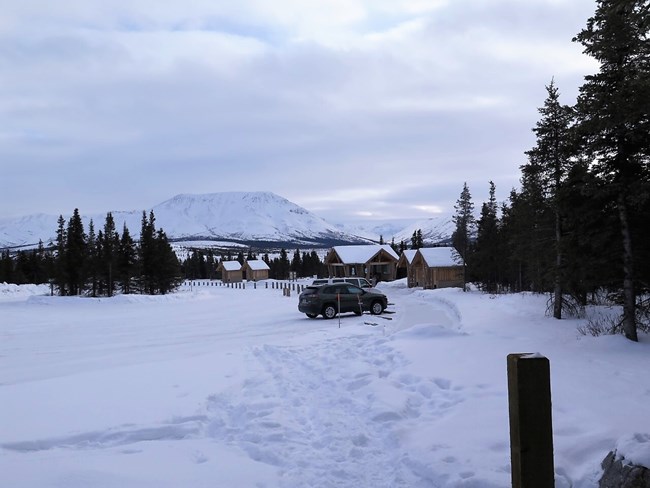 A snow-covered trail leads to a snowy parking lot with a view of mountains in the distance. One car is parked next to a few small wooden outhouses.