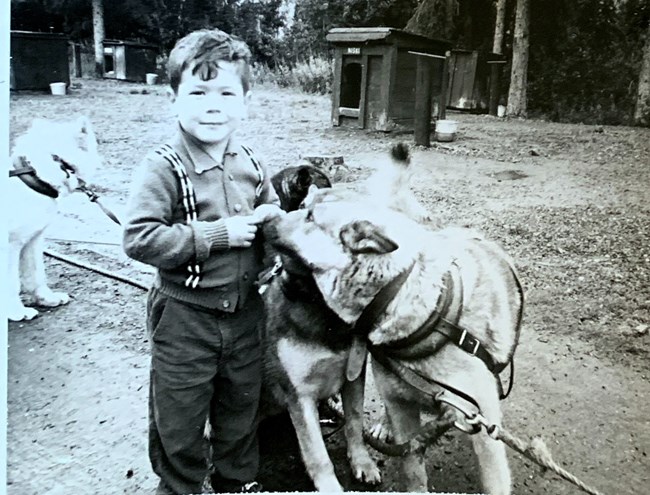 A young boy stands next to a sled dog in a black and white photo