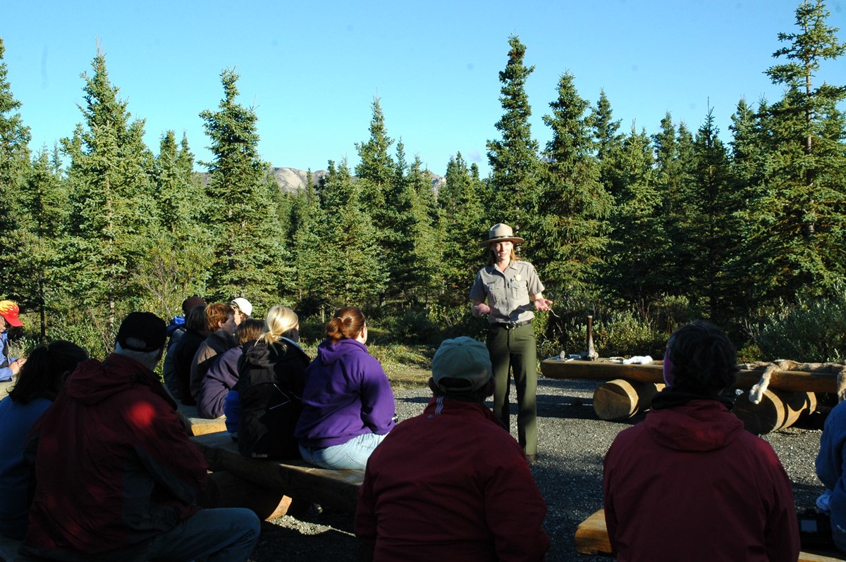 A ranger stands and speaks in front of a crowd of visitors sitting outdoors on wooden benches.