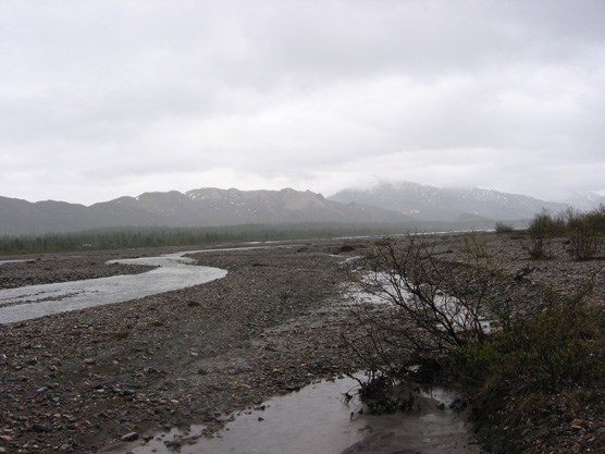 a wide gravel plain with a narrow river flowing through it, mist-shrouded mountains in the distance