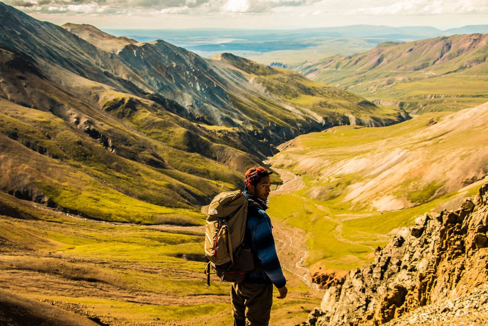 a man with a backpack looking out from a mountainside at a creek far below