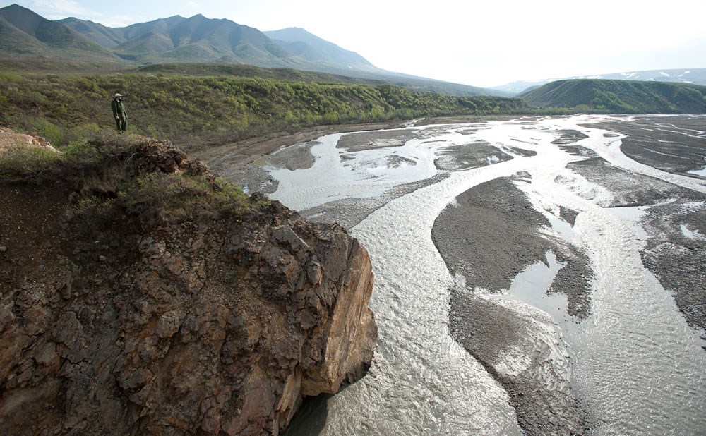 a man standing on a large rocky outcropping, overlooking a braided river