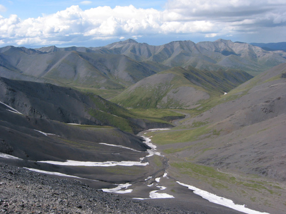 a rocky, snow-dotted slope leading down to a creek, mountains in the distance
