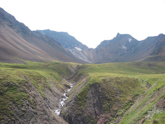 steep, rocky mountains sloping down to a green, fairly level plain, with a narrow creek cutting a deep ditch through its middle