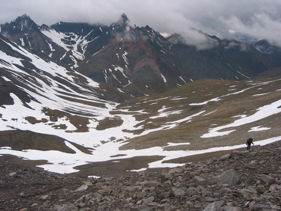 mountains covered at the top by thick clouds, with patches of snow and a person, dwarfed in the scenery, hiking among rocks
