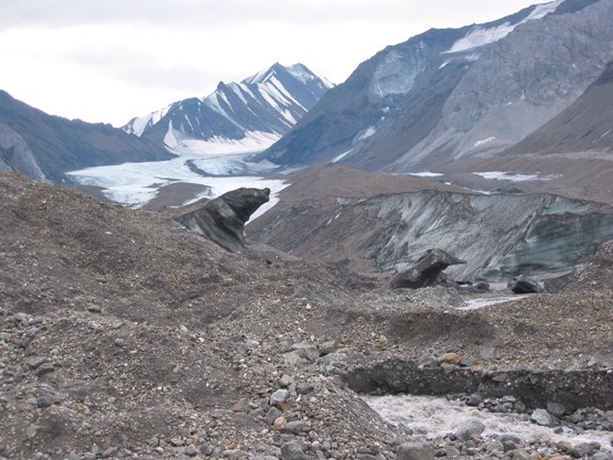 a rocky, uneven landscape leading up to a small glacier and steep, snow-dotted mountains