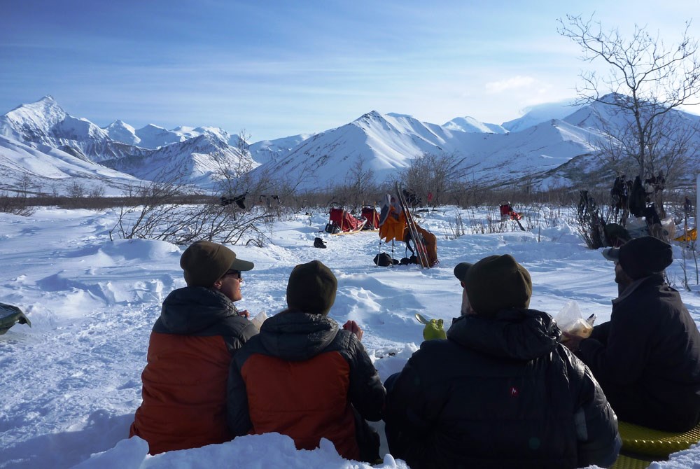 people in winter attire sitting on the snow, looking at a nearby tent and distant, snowy mountains