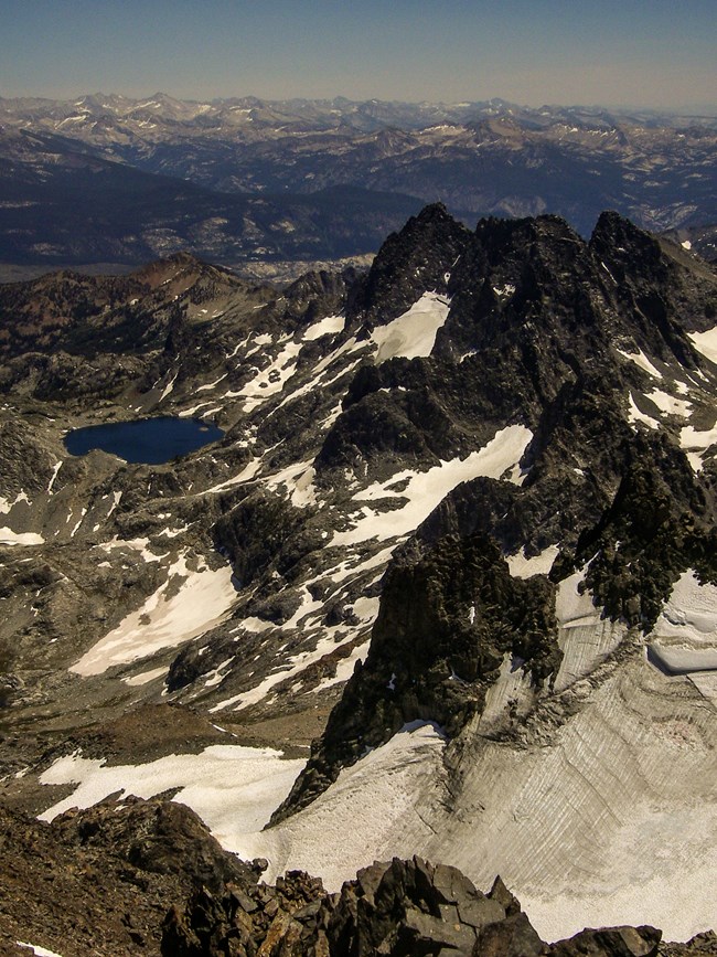 View from a mountain peak looking down on a series of lower peaks to the right, with a small round lake to the left and more mountain peaks in the distance