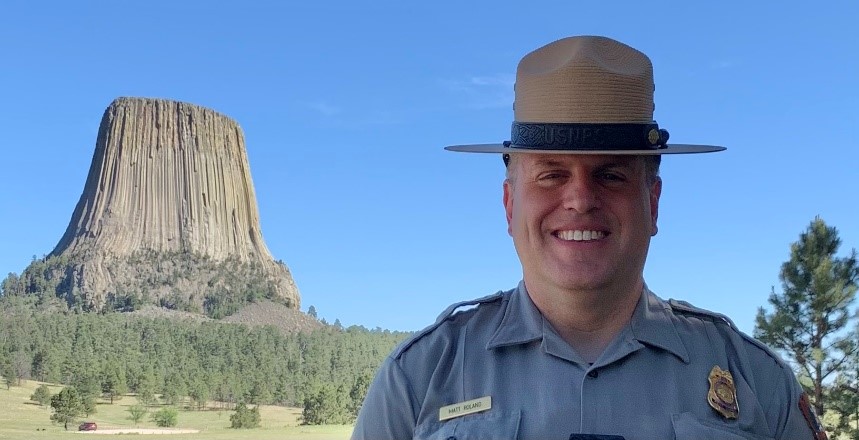 Chief Ranger Matt Roland a man in a ranger uniform smiles in front of Devils Tower, a tall dark gray rock tower formation