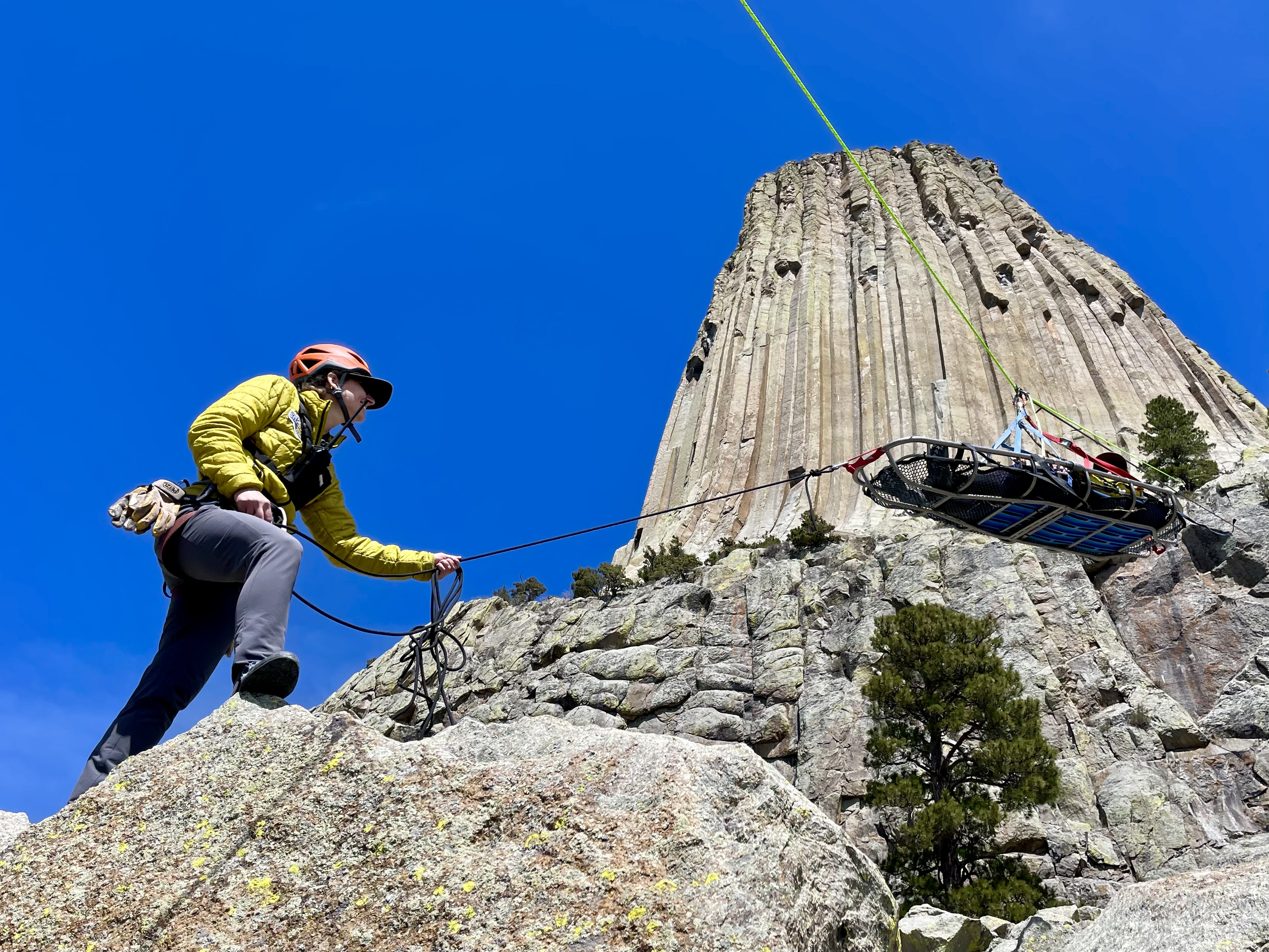 Person in yellowjacket holding rope with litter attached with devils tower in background on a blue sky day