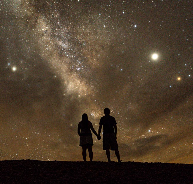 Dark silhouette from behind of a standing man and woman holding hand with a dark sky full of stars and the Milky Way infront of them.