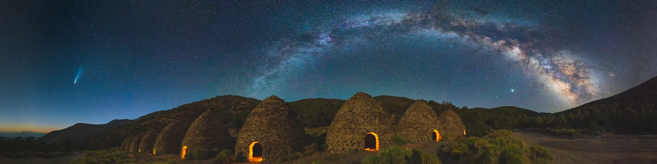 Kilns Pano Panorama of ten beehive shaped masonry charcoal kiln structures at night with yellow light shining through the doorways, the milkyway to the right and the Neowise comet to the left.