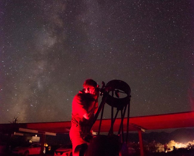 Night image of a person looking through a Dobsonian telescope with a car shade structure in the background and stars above.