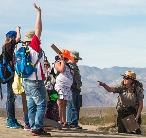 Kneeling on the sand dunes, a ranger points towards students raising their hands.