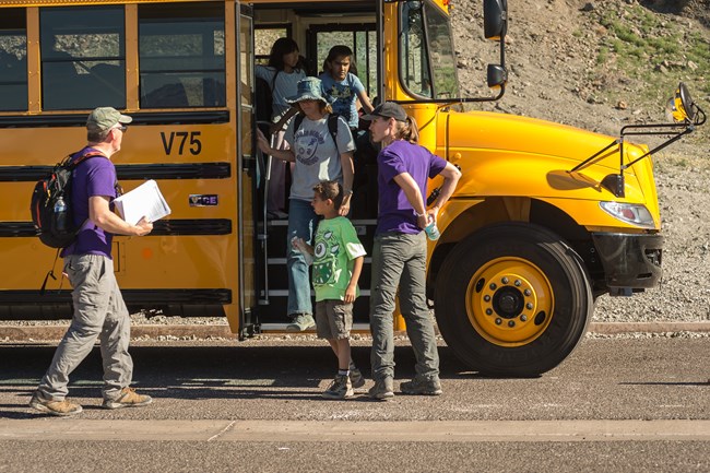 Students and teachers getting off a yellow school bus with a brown rocky natural wall in the background.