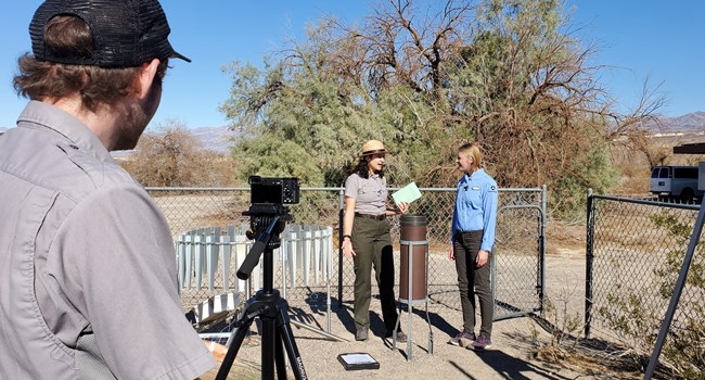 A park ranger operates camera equipment to film two rangers at a weathering station