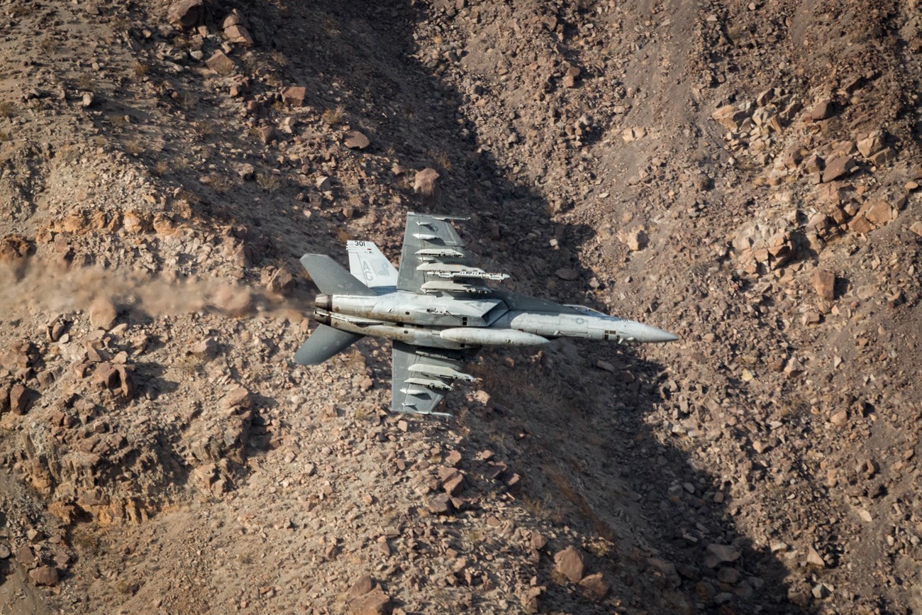 A fighter jet flies through a desert canyon.