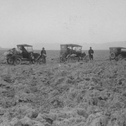 Black and white image of three old vehicles crossing jagged salt flats with people posing for the camera near and on the vehicles.