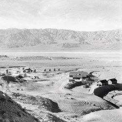 Black and white image of several buildings in a barren desert landscape with mountains in the distance.
