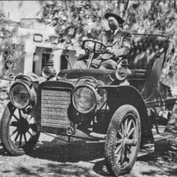 Black and white image of a man driving an old car with part of a building and tree in the background.