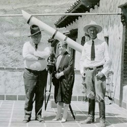 Black and white image of two men and a woman standing on a tile porch with a telescope and part of a building in the background.