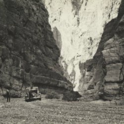 Black and white image of a car and a person in the bottom of a gravel wash with narrow vertical rock walls towering on all sides.
