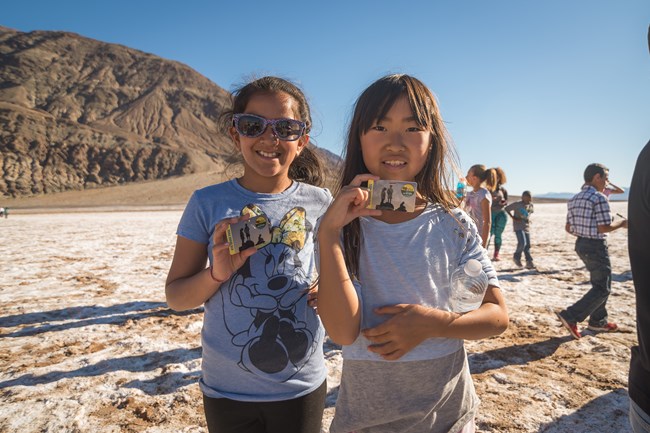 Two girls stand on a salt flat holding Every Kid in a Park passes with additional kids and mountains in the background.