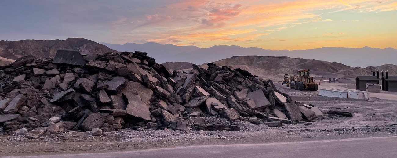 Asphalt piled on the side of the road at Zabriskie Point, yellow dozers to the right work to clear debris under a pink and yellow sunset sky.