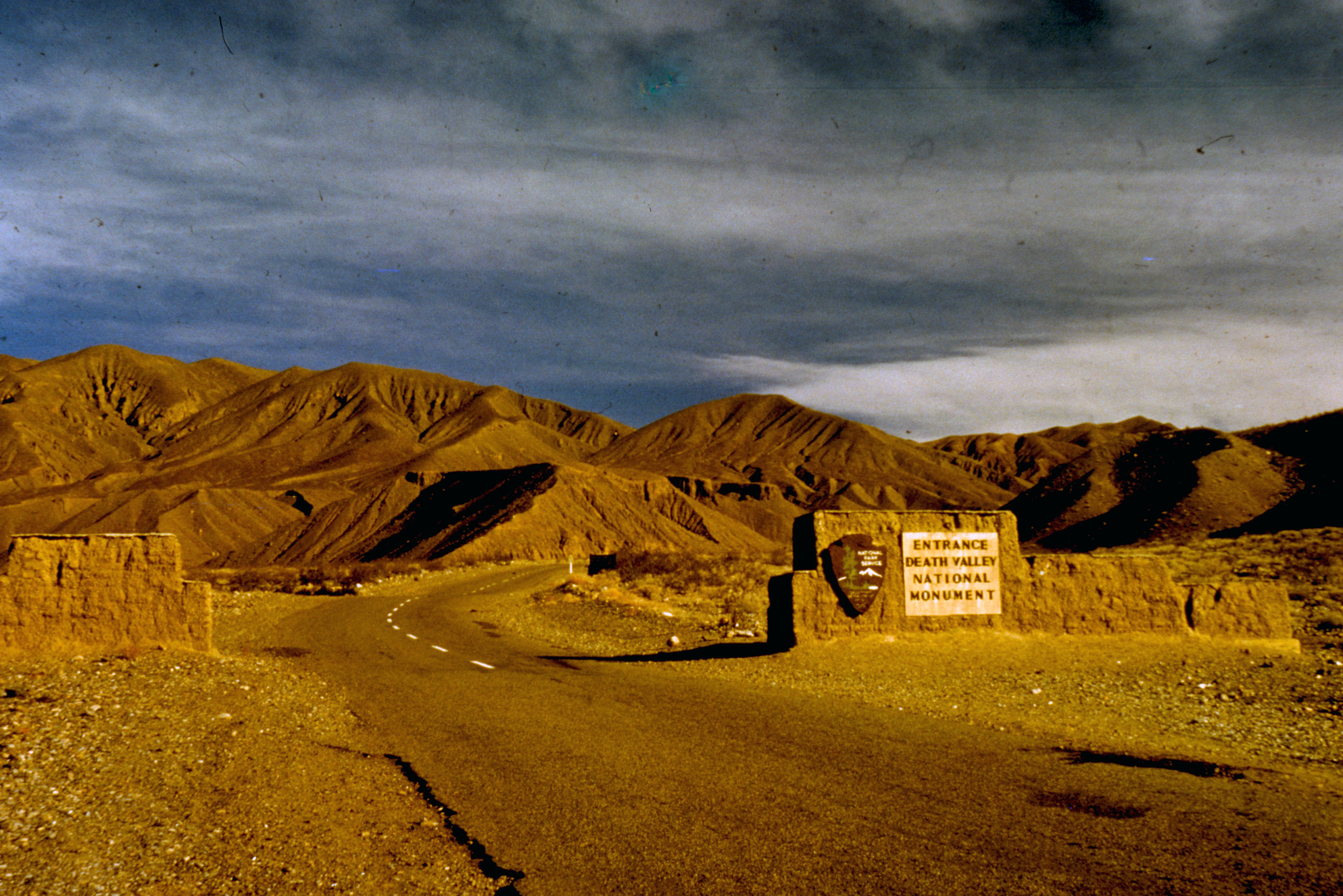 An adobe wall about 5ft tall on both sides of a paved road leading toward desert hills. There is a brown Park Service arrowhead on the wall on the right, with a sign to its right reading "Entrance Death Valley National Monument"