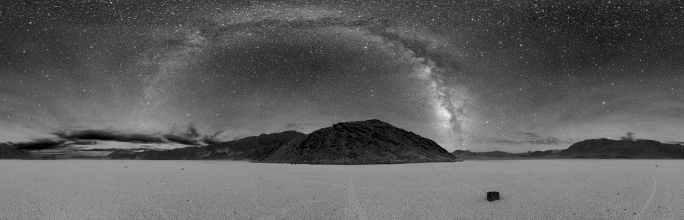 Black and white photograph of the Milky Way arching over desert peaks with a cracked playa and rock with a gouge streak behind it in the foreground.
