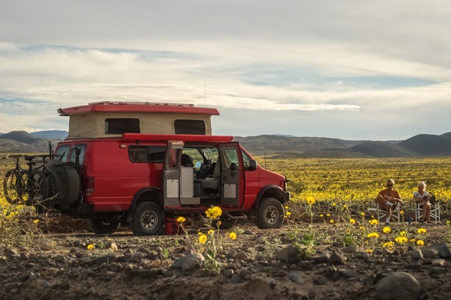 Red camper van on a dirt road surrounded by a field of yellow flowers with two people sitting in camp chairs near the front of the van reading.