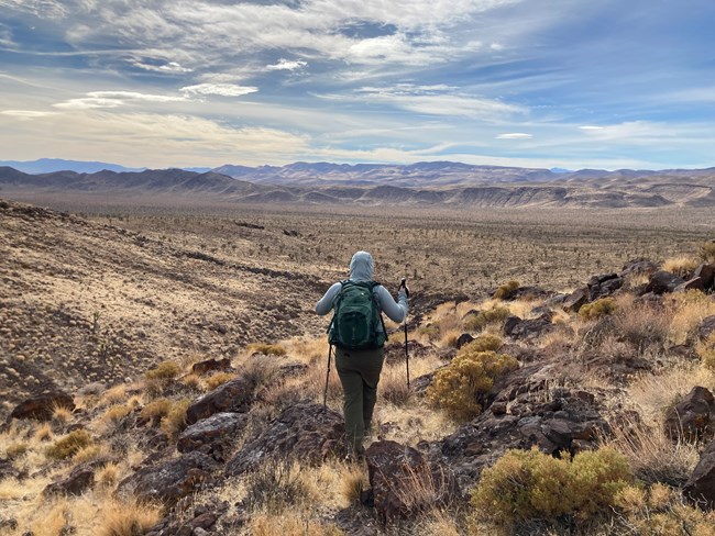 Hiker looks away from the camera toward an open desert landscape with bare mountains and wispy clouds in the distance.