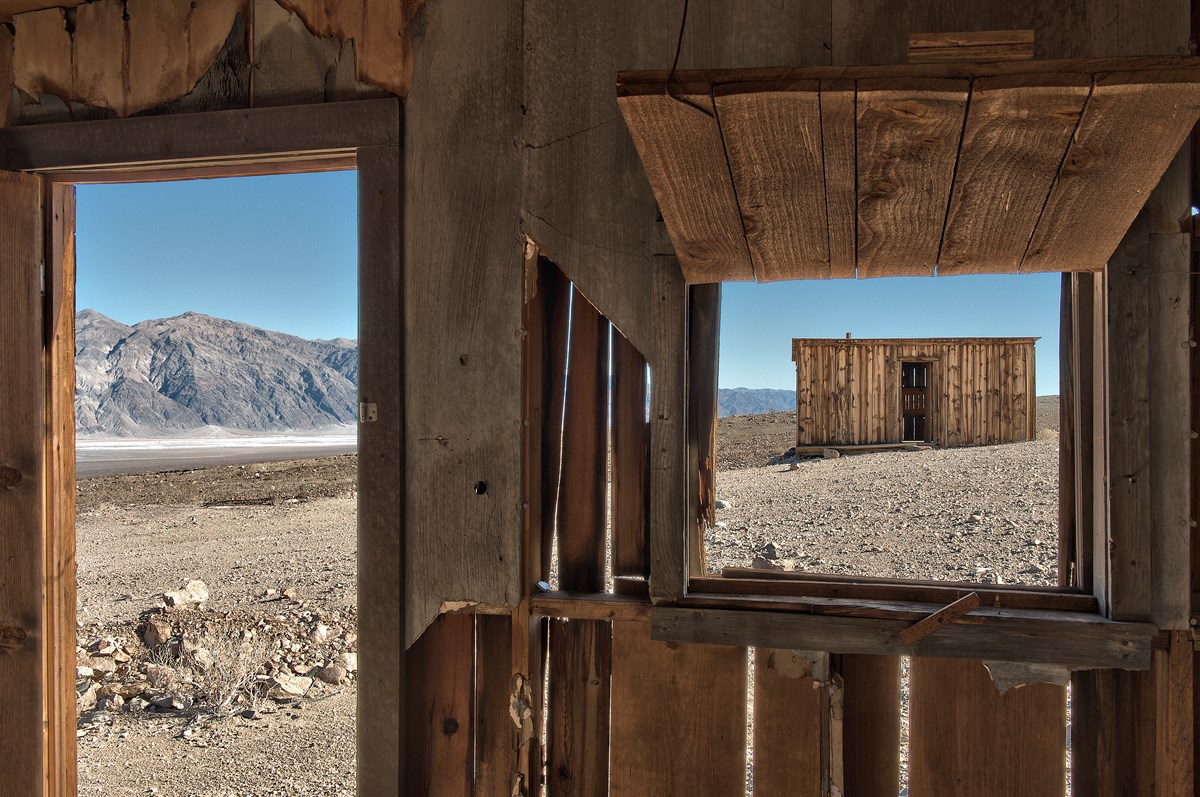 View of a deteriorating wooden cabin from the glassless window of another historic wooden cabin in a bare desert landscape.